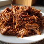 A close-up of tender, saucy crock pot pulled pork piled high on a white plate.