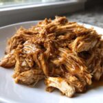Close-up of moist, shredded crockpot teriyaki chicken coated in glossy sauce on a white plate.