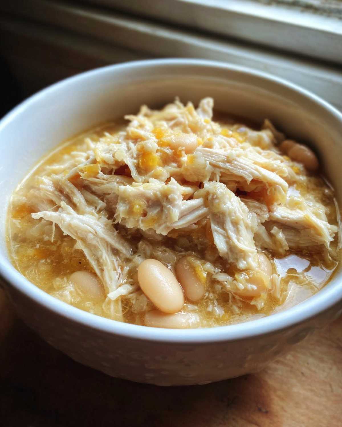 A close-up view of a white bowl filled with rich, shredded chicken and white beans from the crockpot white chicken chili.