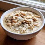 A close-up of a white bowl filled with creamy crockpot white chicken chili, featuring shredded chicken and white beans.