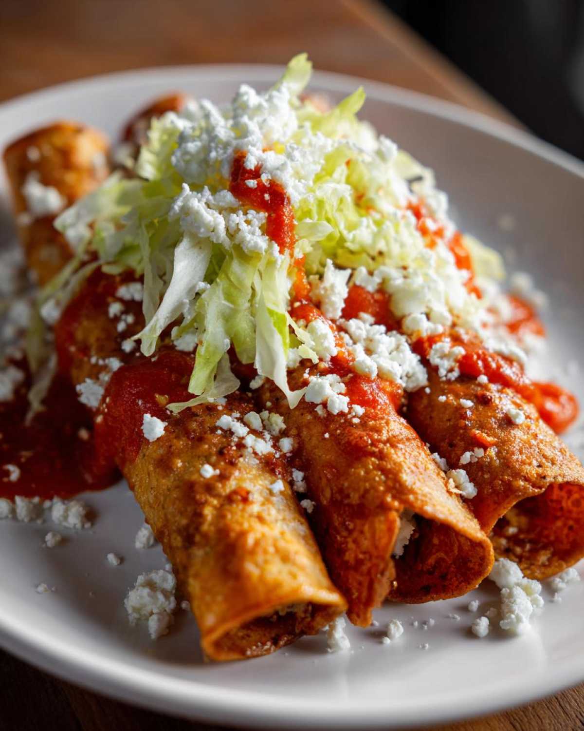 Close-up of three crispy rolled potato tacos smothered in red salsa and topped with shredded lettuce and crumbled white cheese.