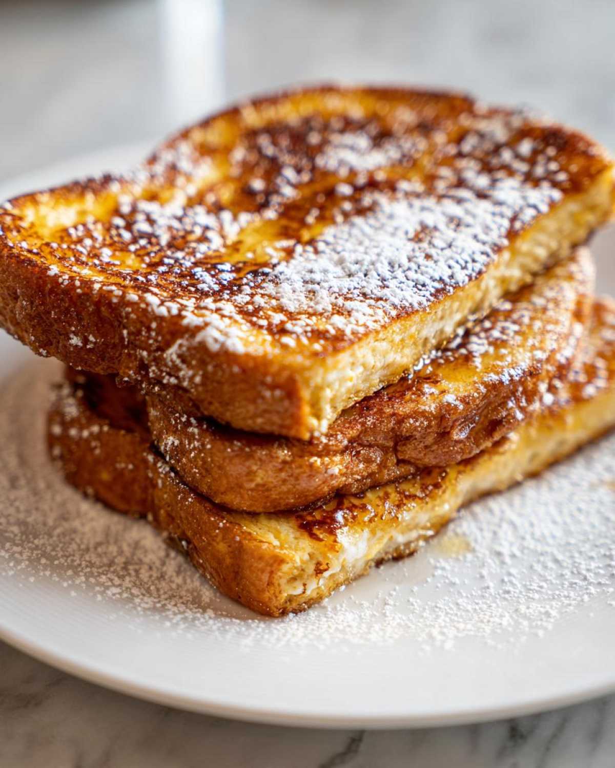 A close-up of three slices of golden brown easy french toast stacked on a white plate, dusted generously with powdered sugar.