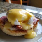 Close-up of a classic eggs benedict featuring a poached egg, ham, and rich hollandaise sauce dripping down.