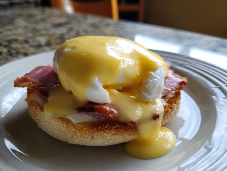 Close-up of a classic eggs benedict featuring a poached egg, ham, and rich hollandaise sauce dripping down.