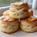 A stack of four golden-brown, flaky buttermilk biscuits drizzled with a light glaze, sitting on a white plate.