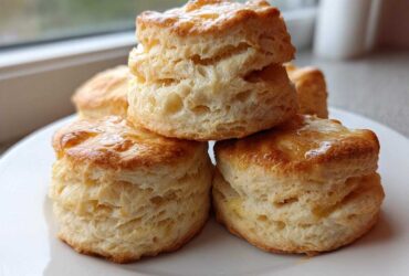 A stack of four golden-brown, flaky buttermilk biscuits drizzled with a light glaze, sitting on a white plate.