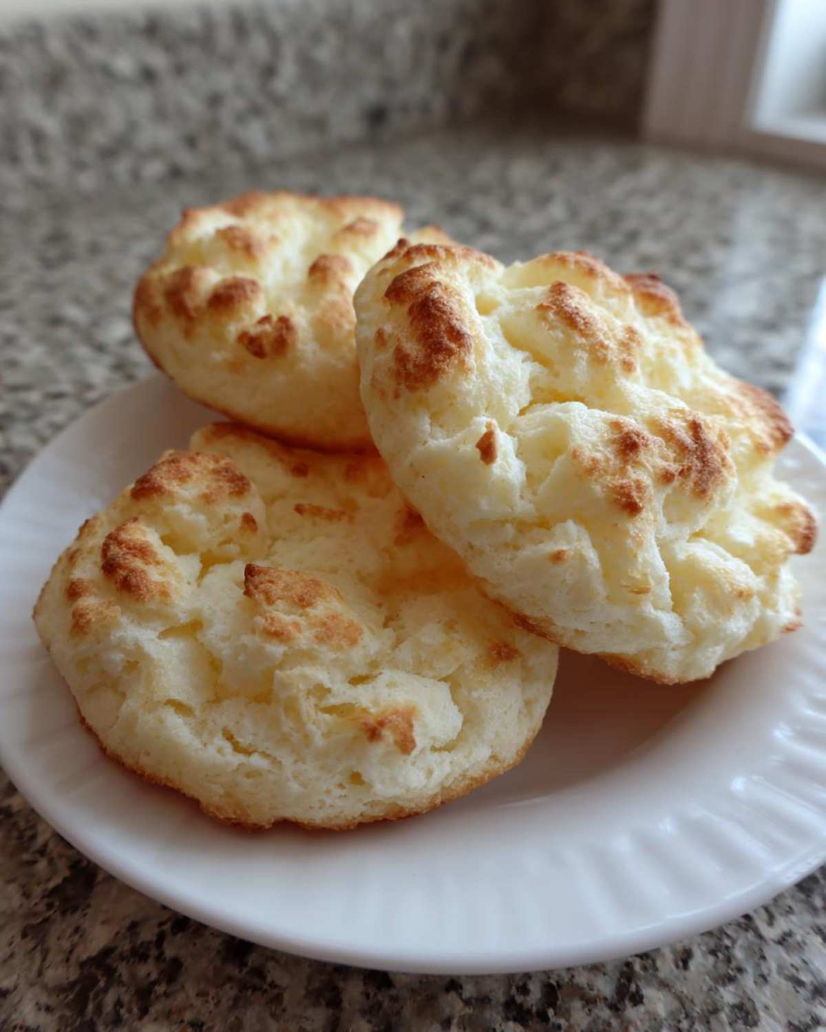 Three golden-brown, fluffy cloud bread pieces resting on a white plate against a granite countertop.