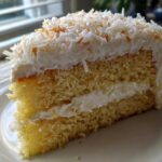 Close-up of a moist slice of layered coconut cake with white frosting and toasted coconut flakes.
