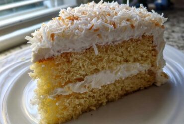 Close-up of a moist slice of layered coconut cake with white frosting and toasted coconut flakes.
