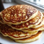 A tall stack of fluffy coconut flour pancakes with golden brown, lacy edges served on a white plate.