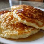 Close-up of fluffy greek yogurt pancakes stacked on a white plate, generously drizzled with syrup.