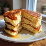 Close-up of a stack of four fluffy pancakes cut in half, showing the airy interior and golden brown tops, drizzled with syrup.