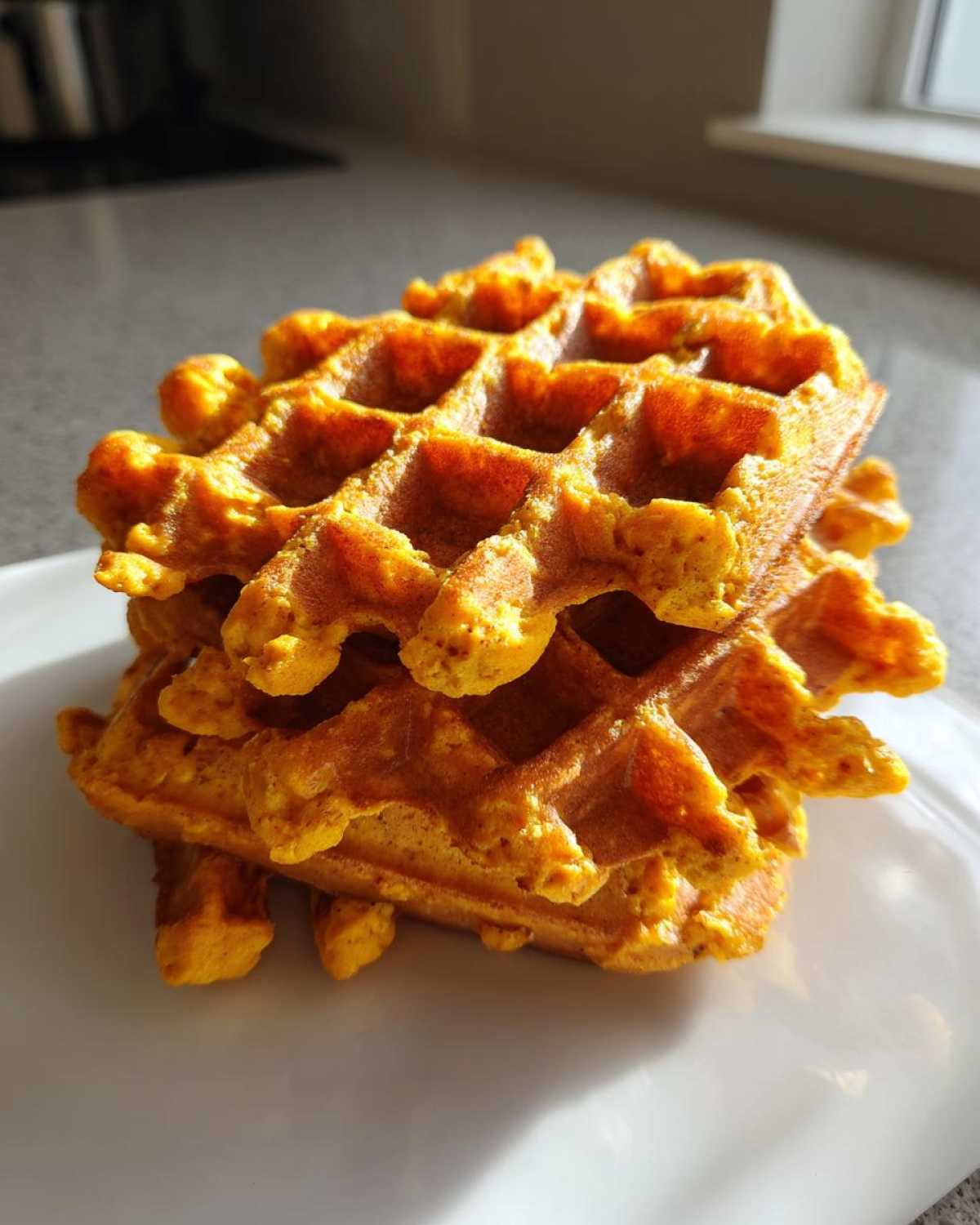 A stack of three golden-orange, fluffy pumpkin waffles resting on a white plate.
