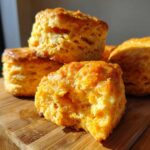 Close-up of fluffy, golden sweet potato biscuits stacked on a wooden board, one broken open to show texture.
