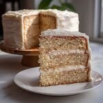 A close-up slice of fluffy vanilla cake recipe layered with white frosting, with the rest of the cake on a stand in the background.