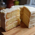 A tall, fluffy vanilla cake recipe with three layers and thick white frosting, a slice cut out.