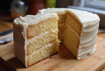 A tall, fluffy vanilla cake recipe with three layers and thick white frosting, a slice cut out.