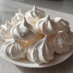 A pile of perfectly piped, bright white meringue cookies displayed on a small white plate.