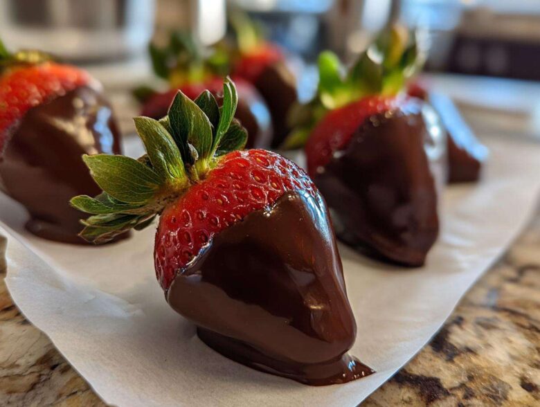 A close-up of fresh chocolate covered strawberries resting on parchment paper, showcasing the glossy dark chocolate coating.