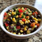 Close-up of fresh cowboy caviar featuring black beans, corn, diced red tomatoes, and cilantro in a white bowl.