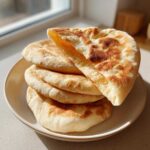 A stack of golden-brown, fluffy homemade pita bread pockets on a light plate, one piece is torn open.