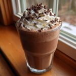 A tall glass of rich, blended frozen hot chocolate topped with whipped cream and chocolate shavings.