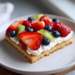 A square slice of fruit pizza sugar cookie topped with whipped cream, strawberries, blueberries, and kiwi.