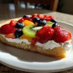 A close-up of a slice of fruit pizza sugar cookie topped with whipped cream, strawberries, blueberries, and kiwi.