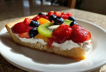 A close-up of a slice of fruit pizza sugar cookie topped with whipped cream, strawberries, blueberries, and kiwi.