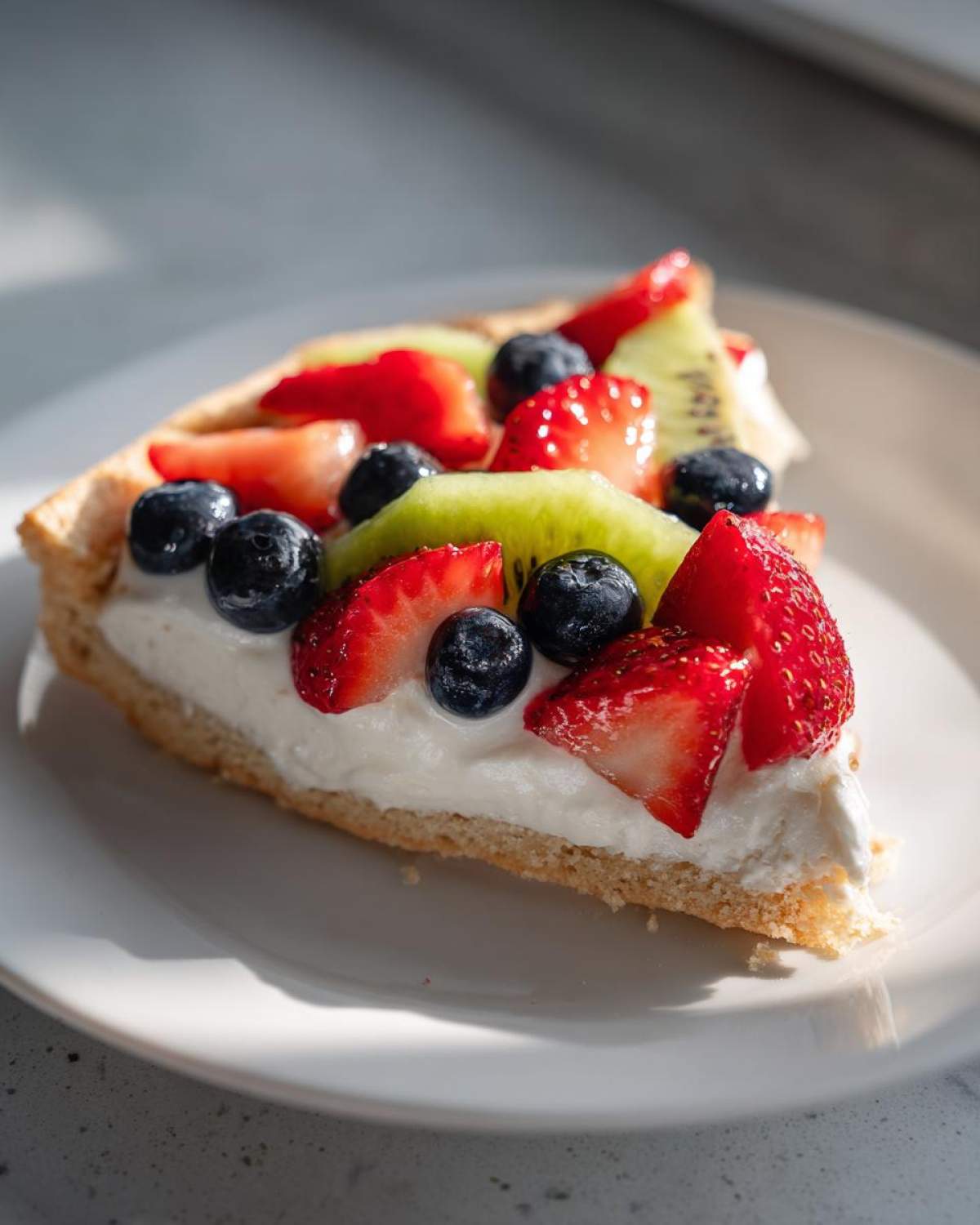 A close-up slice of fruit pizza sugar cookie topped with cream cheese frosting, strawberries, blueberries, and kiwi.