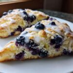 Two triangular blueberry scones, generously studded with fresh blueberries and drizzled with white glaze, resting on a white plate.