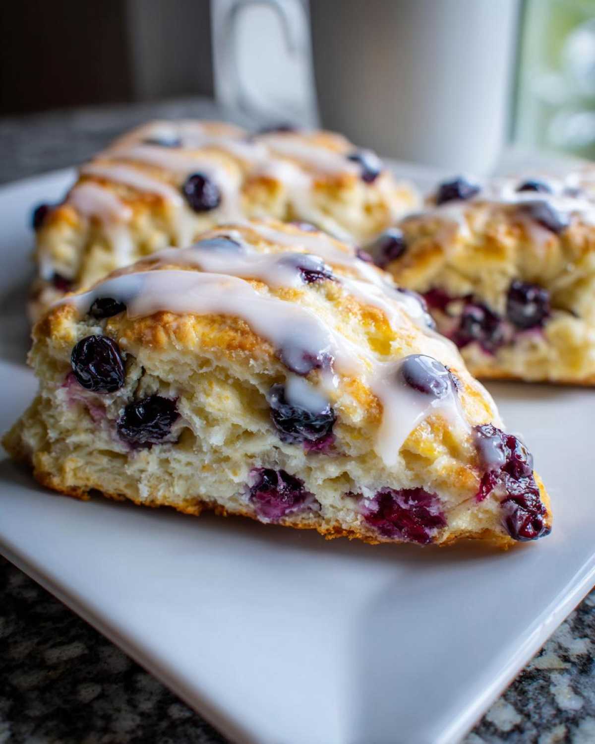 A close-up of a freshly baked, triangle-shaped blueberry scone topped with white glaze, featuring visible blueberries inside.