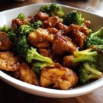 Close-up of saucy, glazed chicken and bright green broccoli florets served together in a white bowl, highlighting the chicken and broccoli dish.