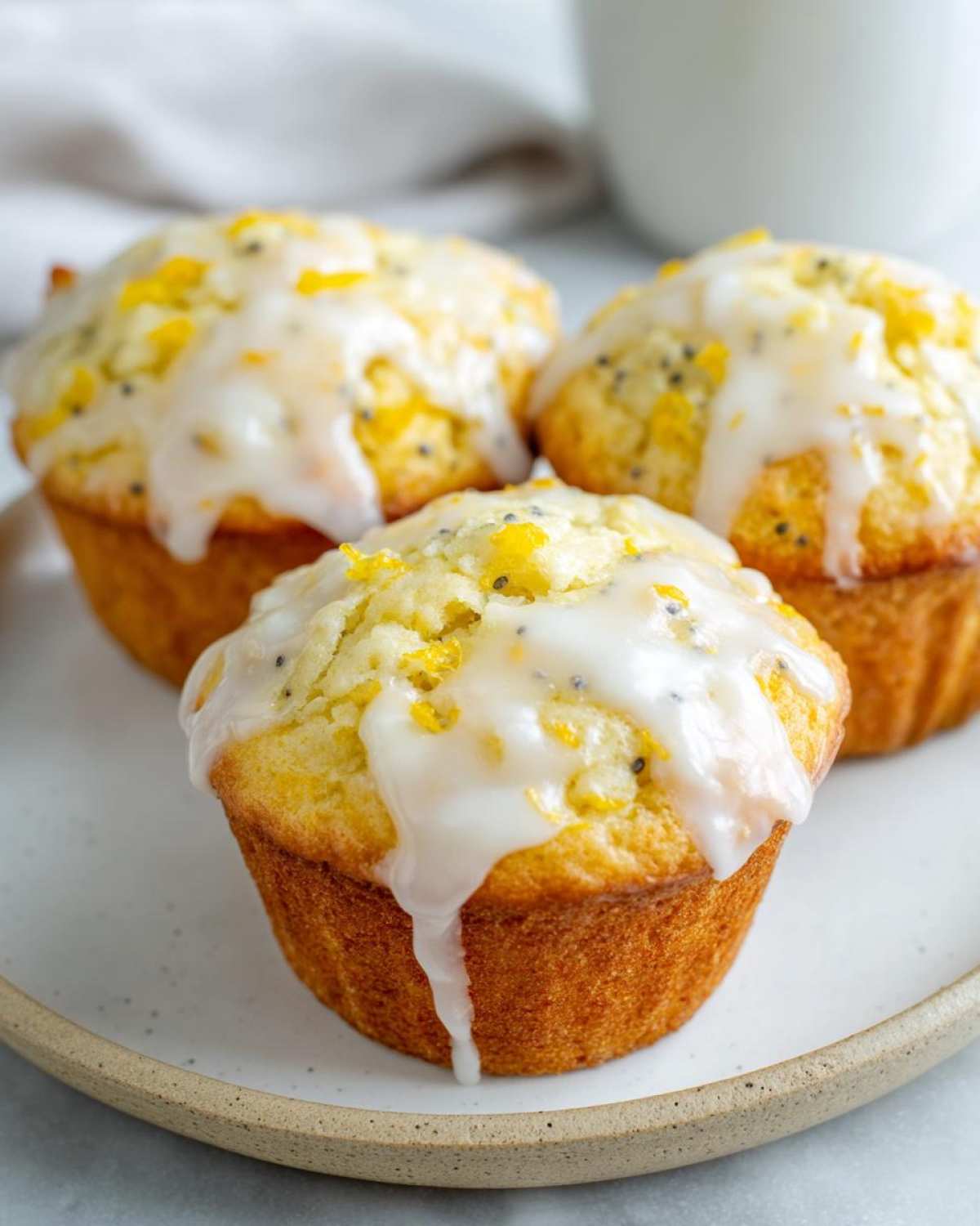 Three golden lemon poppy seed muffins topped with thick white lemon glaze and zest.