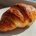 A close-up of a golden brown, flaky baked ham and cheese croissant resting on a white plate.