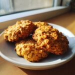 Four golden brown, textured breakfast cookie treats stacked on a light plate near a window.