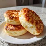 Three golden brown, fluffy rounds of 4-ingredient cloud bread stacked on a white plate in bright sunlight.