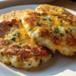 Three crispy, golden brown mashed potato cakes topped with melted cheddar cheese and fresh chives, served on a white plate.