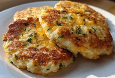 Three crispy, golden brown mashed potato cakes topped with melted cheddar cheese and fresh chives, served on a white plate.