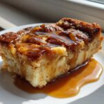 A square slice of gooey bread pudding topped with rich caramel sauce, served on a white plate.