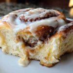 A close-up shot of a soft, freshly baked cinnamon roll topped with dripping white icing, showing the sweet cinnamon swirl inside.