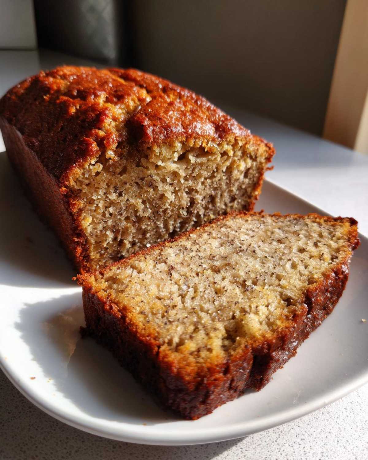 A close-up of a freshly baked greek yogurt banana bread loaf with one slice cut and resting beside it on a white plate.