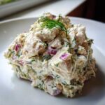 A mound of creamy greek yogurt chicken salad mixed with red onion and fresh dill, served on a white plate.
