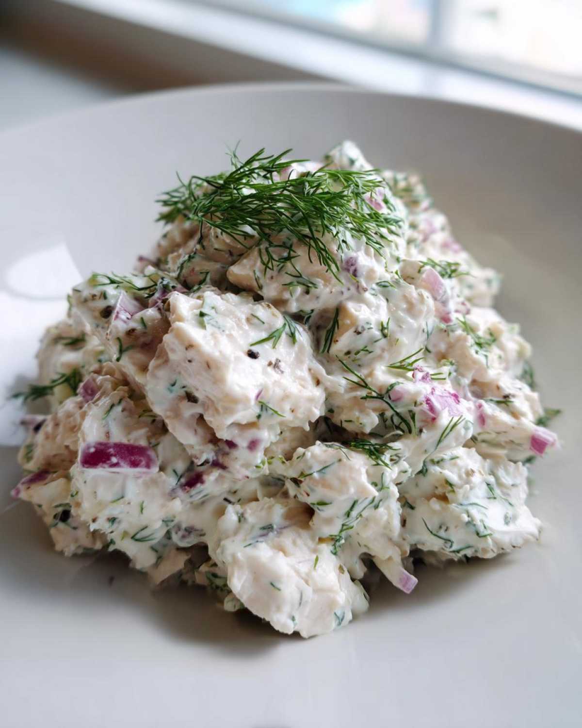 A mound of creamy greek yogurt chicken salad mixed with red onion and fresh dill, served on a white plate.