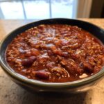 Close-up of a dark bowl filled with rich, thick, hearty chili featuring ground meat and kidney beans.
