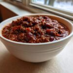 A close-up of a white bowl filled with rich, dark red hearty chili featuring ground meat and beans.