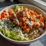 A close-up of a vibrant buffalo chicken bowl featuring shredded orange buffalo chicken, brown rice, blue cheese crumbles, carrots, and ranch dressing.
