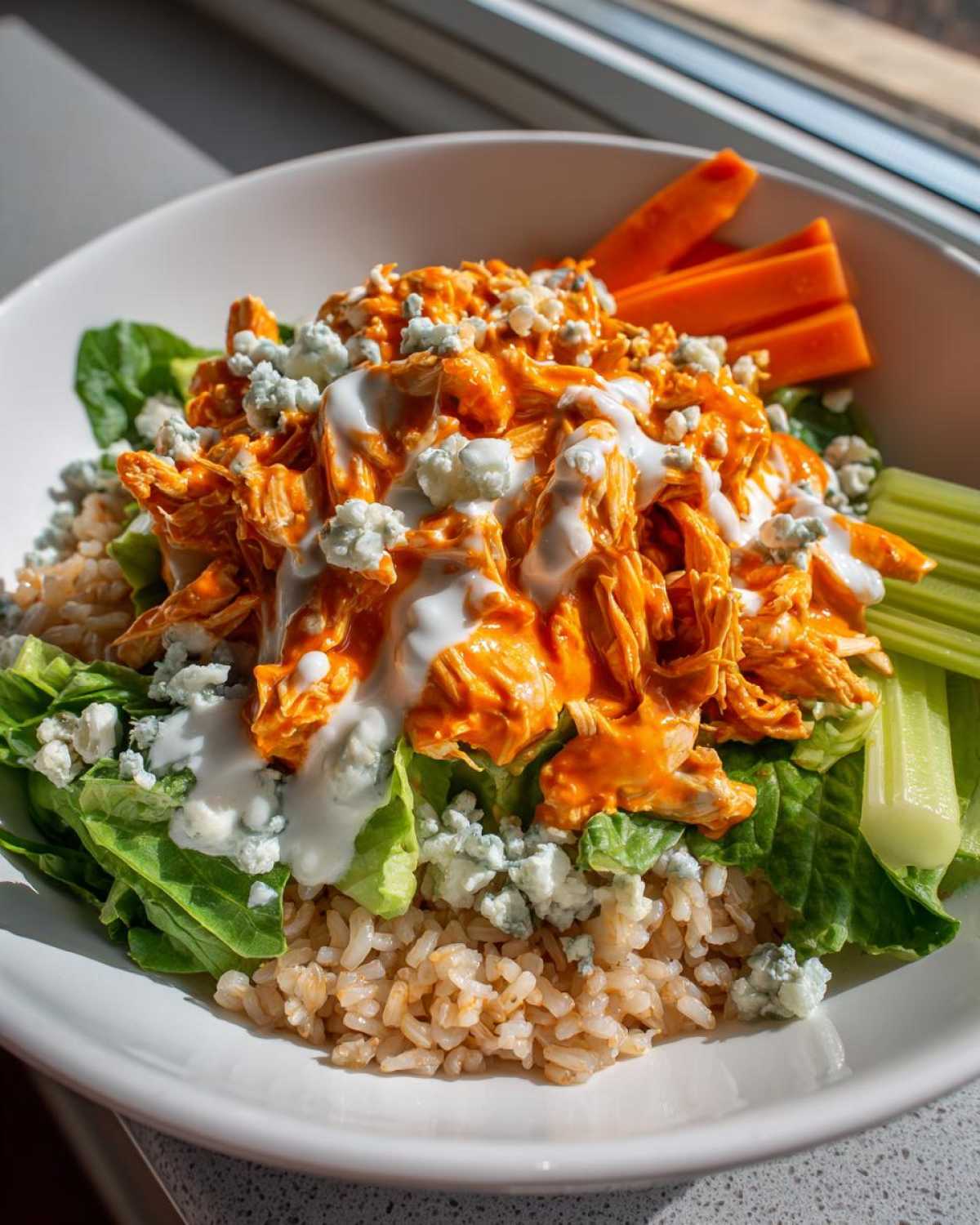 A close-up of a healthy buffalo chicken bowl featuring shredded orange buffalo chicken, brown rice, lettuce, blue cheese crumbles, and ranch dressing.