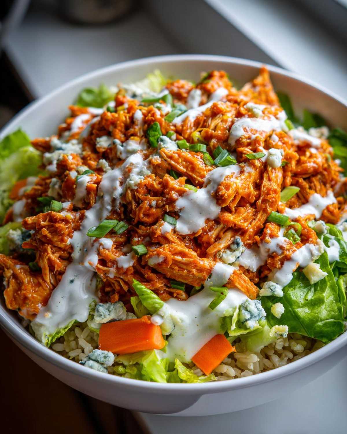 Close-up of a vibrant buffalo chicken bowl featuring shredded orange buffalo chicken, lettuce, brown rice, blue cheese crumbles, and a drizzle of ranch dressing.