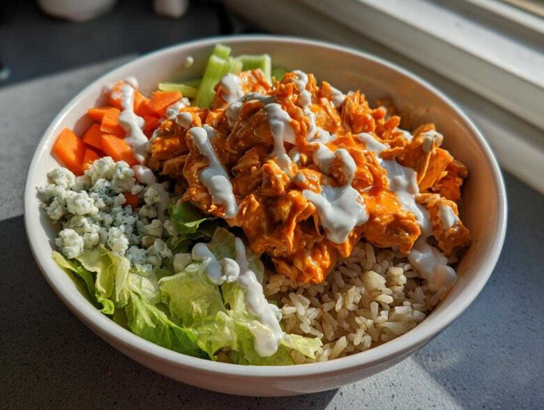 A close-up of a vibrant buffalo chicken bowl featuring shredded orange buffalo chicken, brown rice, blue cheese crumbles, carrots, and ranch dressing.
